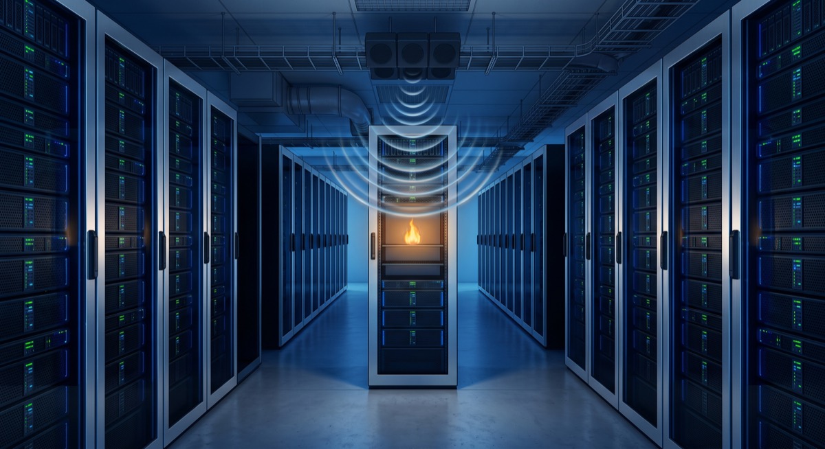 A server room mid-suppression: racks of equipment gleaming, a single small flame in one rack already going out—not a drop of water anywhere, the air around the fire visibly disturbed but everything else dry, clean, and very much still running